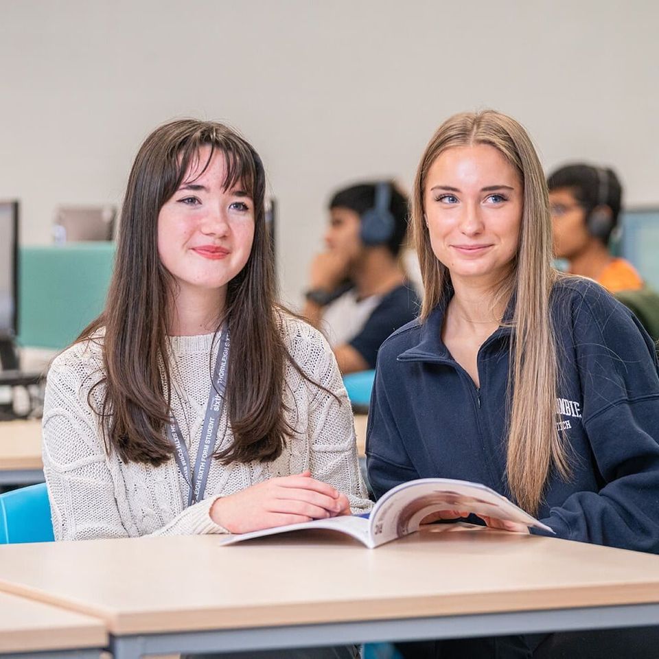 Two people are sitting at a table in a classroom, smiling and looking at a book.